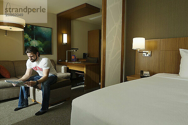 Ein kluger junger Mann sitzt auf einem Hocker in einem Hotelzimmer und liest eine Zeitung.