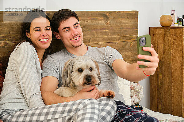 Spanisches glückliches Paar macht ein Selfie mit seinem Hund im Bett.