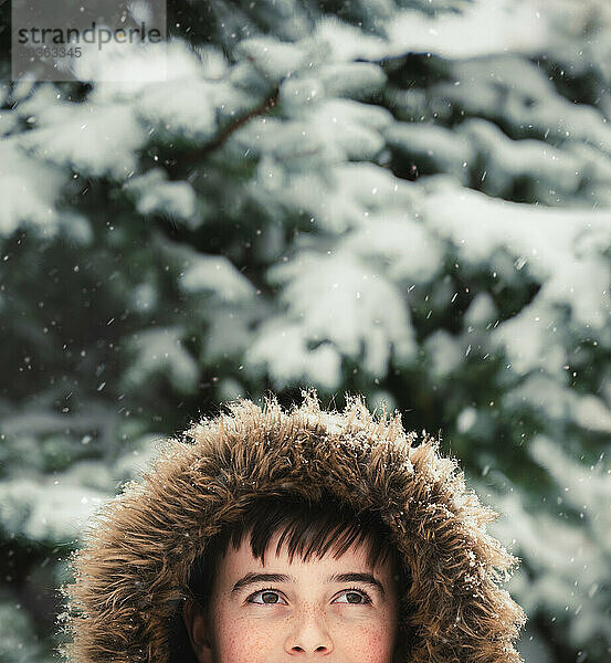 Glücklicher Junge trägt Mantel mit pelziger Kapuze draußen im Schnee an einem Wintertag.
