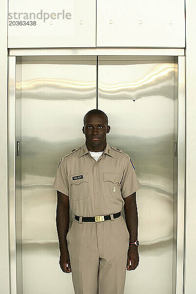 A young handsome African American male stands firm in front of an elevator wearing his uniform.