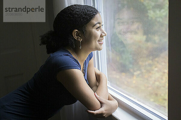 Ein lächelndes gemischtrassiges Teenager-Mädchen blickt aus einem Fenster