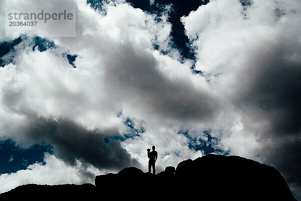 Breite Silhouette eines Vaters  der sein Kind auf einem Hügel mit Wolken hält.