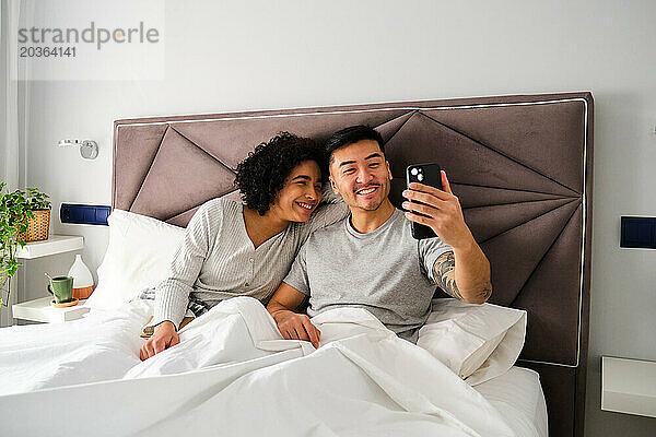 Multiethnisches Paar lächelt und macht ein Selfie im Bett.