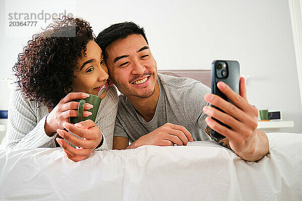 Gemischtrassiges Paar lächelt und macht ein Selfie im Bett.