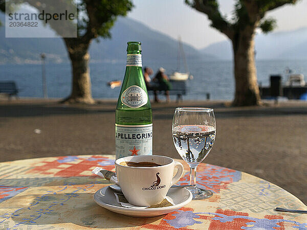 Eine Tasse Kaffee und eine Flasche Mineralwasser mit Kohlensäure auf dem Tisch einer Bar am Boulevard von Ascona  einer Stadt im Schweizer Tessin. Im Hintergrund ist der Lago Maggiore zu sehen.