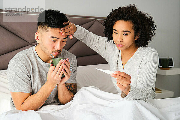 Eine lateinamerikanische Frau misst das Fieber seines asiatischen Mannes mit einem Thermometer.