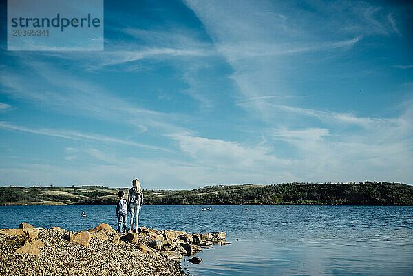 Weite Rückansicht eines Teenager-Mädchens und eines kleinen Jungen am felsigen Ufer des Sees.