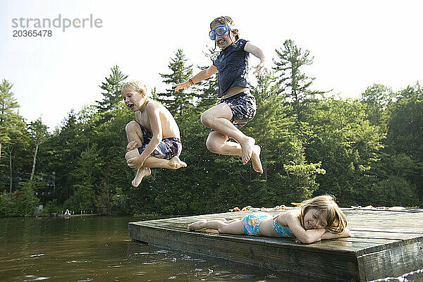 Zwei Kinder springen von einem Holzfloß in Center Harbor  New Hampshire  über einen Thrid in einen See.