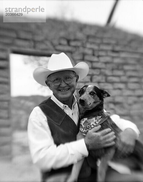 Ein älterer Herr posiert mit seinem kleinen Hund vor einer alten Ranch gegen eine Umfrage.
