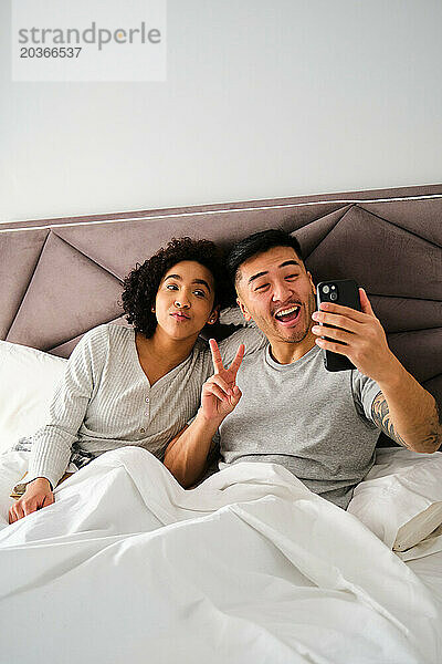 Vielfältiges Paar lächelt und macht ein Selfie im Bett.