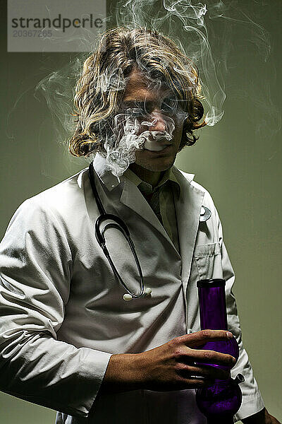 Ein als Arzt verkleideter Mann atmet Rauch aus  während er eine rauchende Bong in der Hand hält.