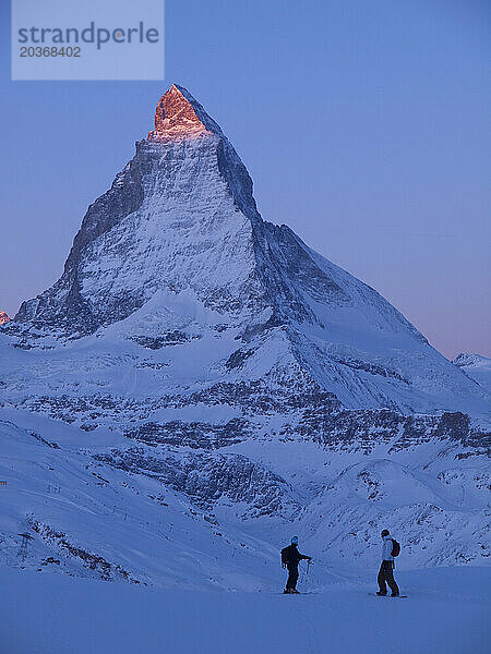 Ein Skifahrer und ein Snowboarder starren auf das Matterhorn  das im ersten Sonnenlicht (Alpenglühen) badet.