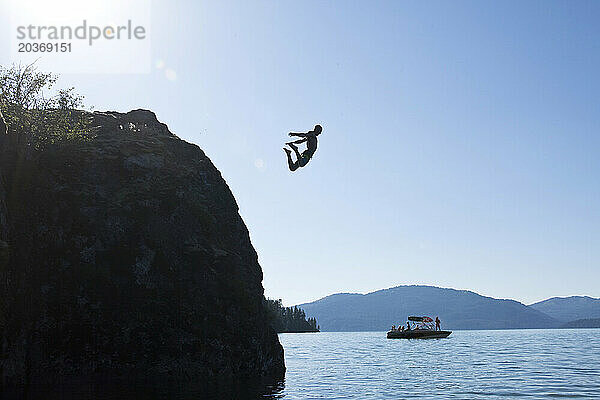 Ein junger Mann springt in einen See mit einem Wakeboard-Boot im Hintergrund in Idaho.