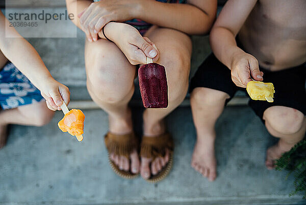 Draufsicht auf drei Kinder mit Eis am Stiel  die draußen sitzen