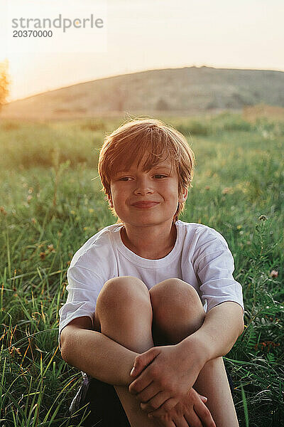 Fröhlicher Junge sitzt auf einer Wiese und trägt ein weißes T-Shirt