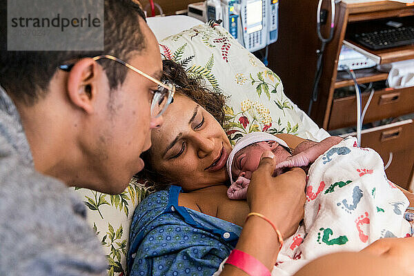 Gemischtrassiges Paar kuschelt sein Neugeborenes direkt nach der Geburt im Krankenhaus