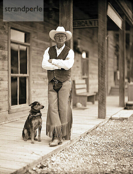 Ein älterer Herr posiert mit seinem kleinen Hund vor einer alten Ranch gegen eine Stange.