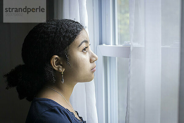 Ein gemischtrassiges Teenager-Mädchen schaut mit ernster Miene aus dem Fenster