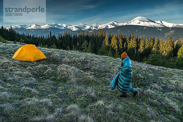 Junge Frau  in eine Decke gehüllt  geht bei Sonnenaufgang auf das Zelt zu
