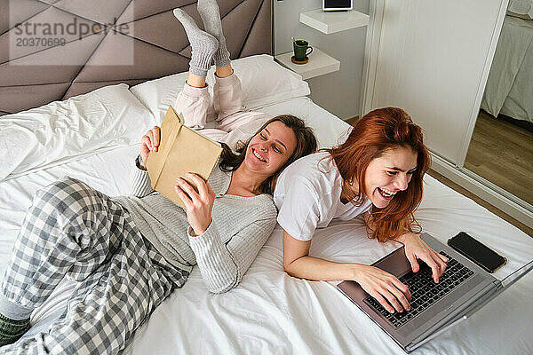 Lesbisches Paar lacht  während es den Laptop benutzt und im Bett ein Buch liest.