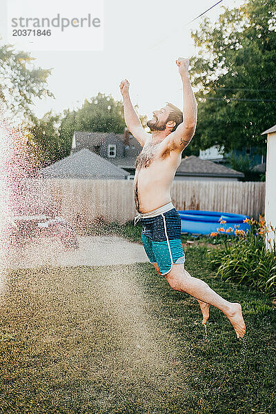 Lächelnder Mann springt im Sommer draußen durch sprühendes Schlauchwasser