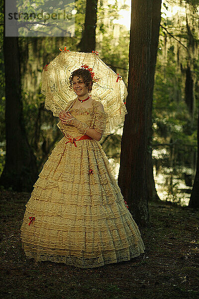 Eine junge Frau posiert in einem Southern Belle-Outfit  einschließlich eines großen  verzierten historischen Kleides und Sonnenschirms  in der Nähe eines Sees.