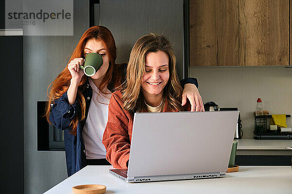 Lesbisches Paar benutzt den Laptop  während es in der Küche Kaffee trinkt.