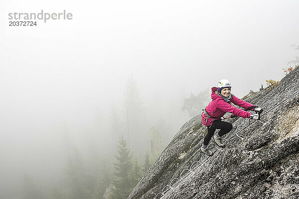 Eine Frau lächelt vor Aufregung  als sie die Metallsprossen eines Klettersteigs in Squamish hinaufklettert.