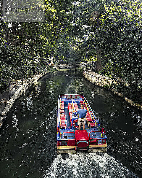 An einem sonnigen Spätsommertag fährt ein Mann mit einem Wassertaxi den San Antonio River Walk entlang. Der Flussweg ist mit einem Blätterdach aus Bäumen bedeckt und bietet einen schönen  ruhigen Ort zum Spazierengehen.