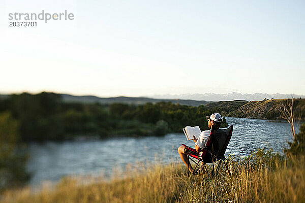 Ein sportlicher Mann liest bei Sonnenuntergang ein Buch  während er auf einer Raftingtour in Montana an einem Fluss campt.