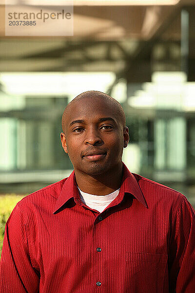 Portrait of a young African American male wearing a red shirt with a blurry modern building in the background.