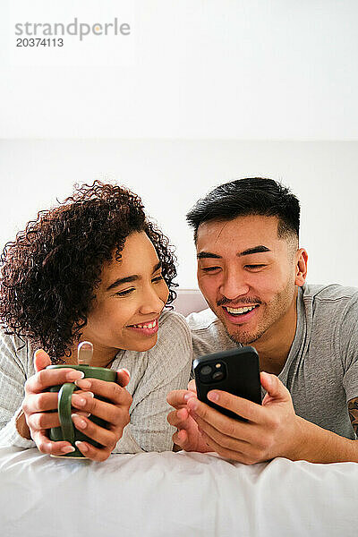 Multiethnisches glückliches Paar nutzt das Smartphone im Bett.