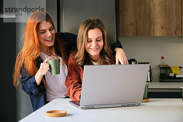 Lesbisches Paar benutzt den Laptop  während es in der Küche Kaffee trinkt.