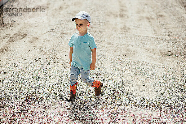 Weite Sicht auf einen kleinen Jungen mit Hut und Stiefeln  der auf einer Schotterstraße läuft