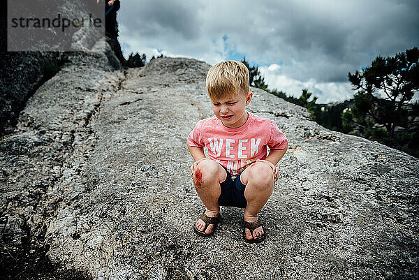 Weite Sicht auf einen kleinen Jungen  der auf Felsen sitzt und mit aufgeschürftem Knie weint.