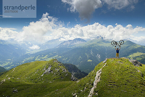 Ein Mountainbiker steht auf einem Gipfel und hält sein Fahrrad in der Luft. Mountainbiken in Lenzerheide. Die Region Graubünden in der Schweiz ist ein Singletrail-Paradies.