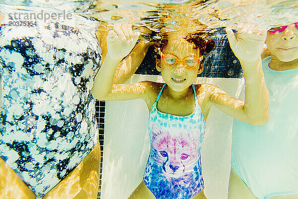 Ein kleines Mädchen trägt eine Schwimmbrille und taucht unter Wasser  ein kleiner Junge und ein weiteres kleines Mädchen auf beiden Seiten.