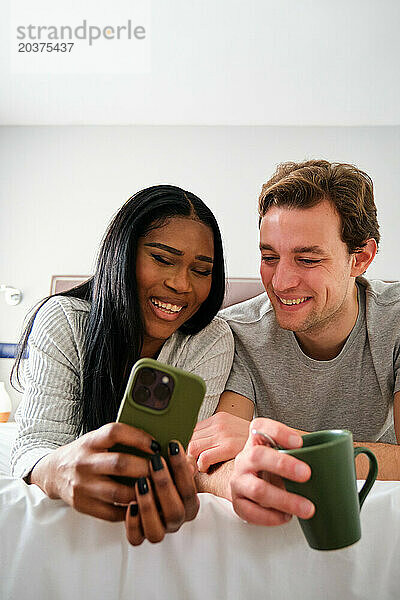Multiethnisches Paar lacht  während es sich Videos auf dem Smartphone ansieht.