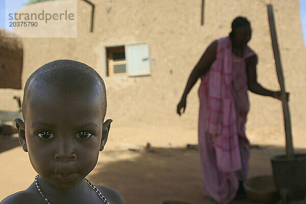 Ein junges afrikanisches Toureg-Mädchen blickt in die Kamera  während ihre Mutter im Hintergrund Hirse und Sorghum zerstampft  Gossi  Mali  Westafrika