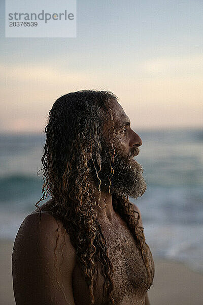 Porträt eines glücklichen Mannes mit langen lockigen Haaren bei Sonnenuntergang am Strand.