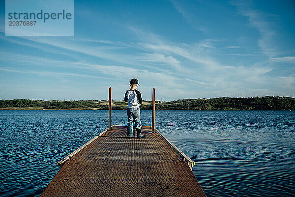 Weite Rückansicht eines kleinen Jungen  der auf dem Dock steht und auf den See blickt.