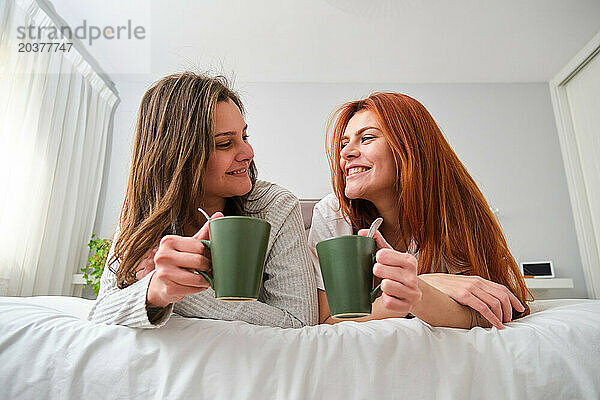 Lesbisches Paar trinkt Kaffee im Bett und schaut einander lächelnd an.