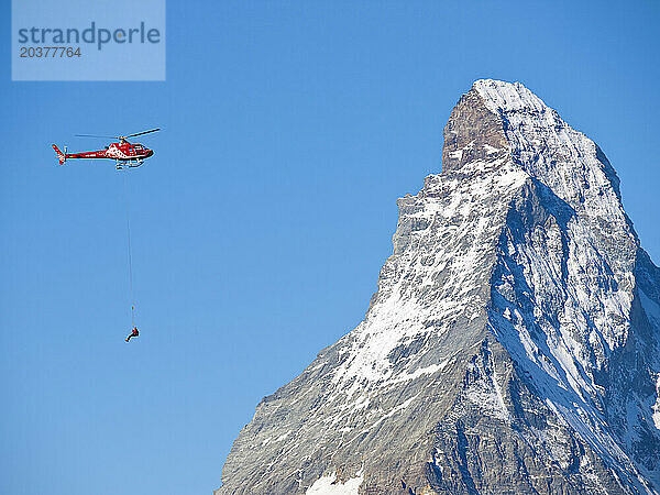 Ein Bergrettungstechniker baumelt während einer Rettungsmission für einen verletzten Bergsteiger in den Schweizer Alpen unter einem Hubschrauber. Im Hintergrund ist der berühmte Berg Matterhorn zu sehen.