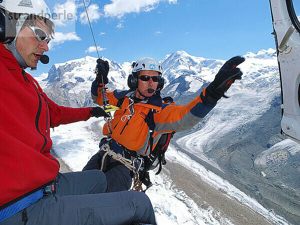 Ein Bergrettungstechniker steigt hoch über dem Boden aus einem Hubschrauber. Ein Windenmann ist bereit  ihn zum Gletscher hinunterzulassen  wo ein Bergsteiger bei einem Sturz verletzt wird.