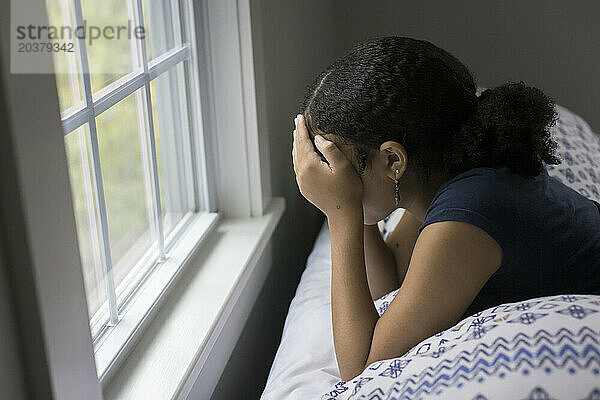 Ein gemischtrassiges Teenager-Mädchen liegt auf dem Bett  den Kopf in die Hände gestützt  und wirkt deprimiert