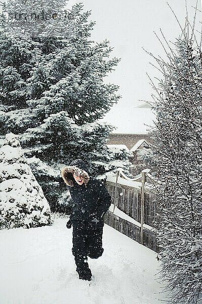 Glücklicher Junge trägt Mantel mit pelziger Kapuze draußen im Schnee an einem Wintertag.