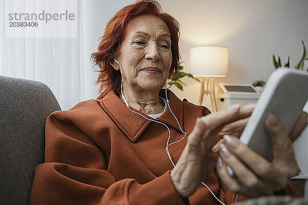 Retired senior woman using smart phone with headphones sitting on sofa in living room