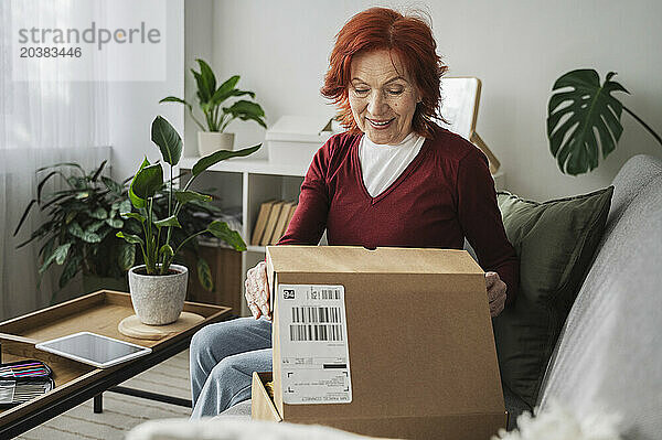 Smiling retired senior woman opening package on sofa at home