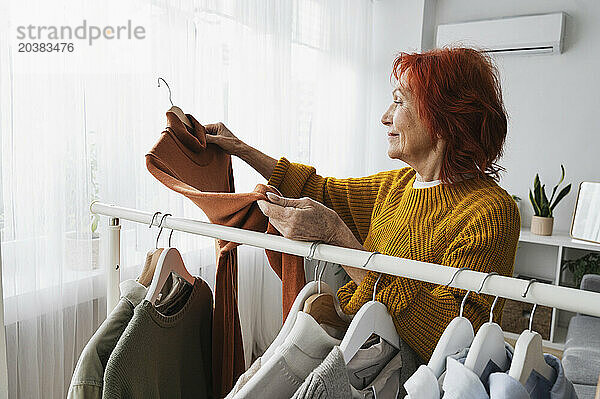 Retired redhead senior woman choosing sweater from clothes rack at home