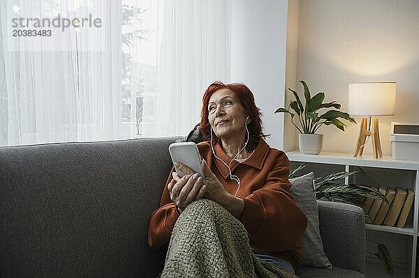 Thoughtful senior woman with smart phone and headphones sitting on couch in living room